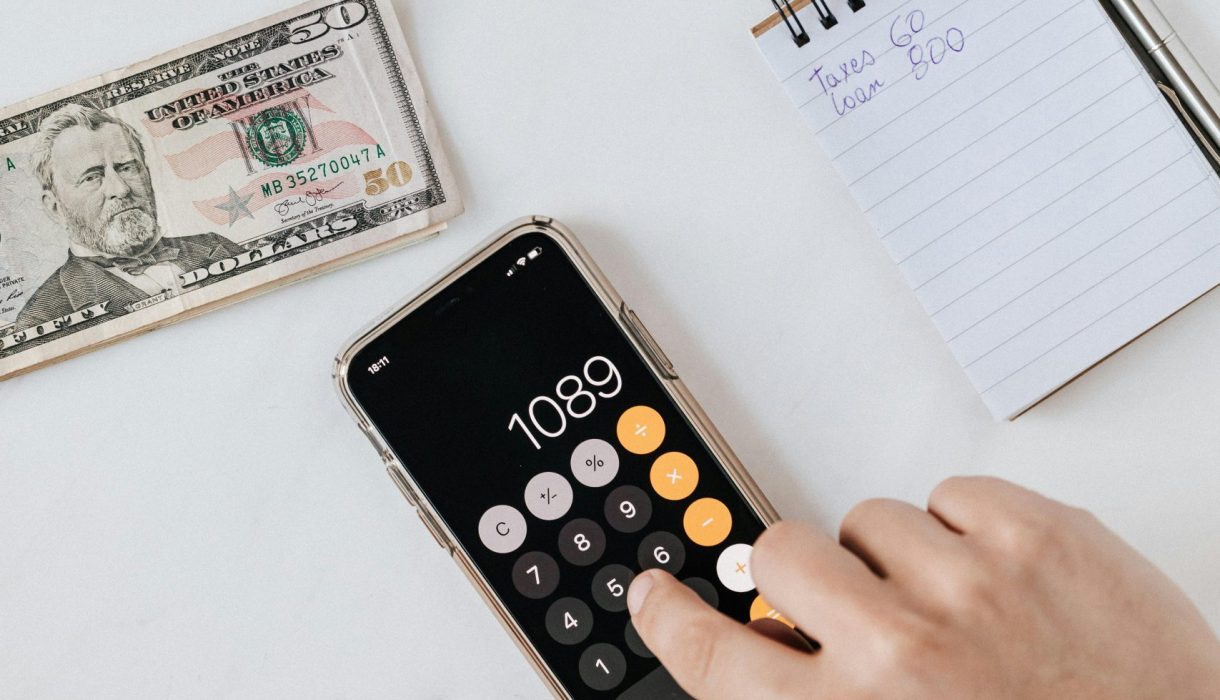 Overhead view of a hand using a calculator with US dollars and notes, representing finance and budgeting.