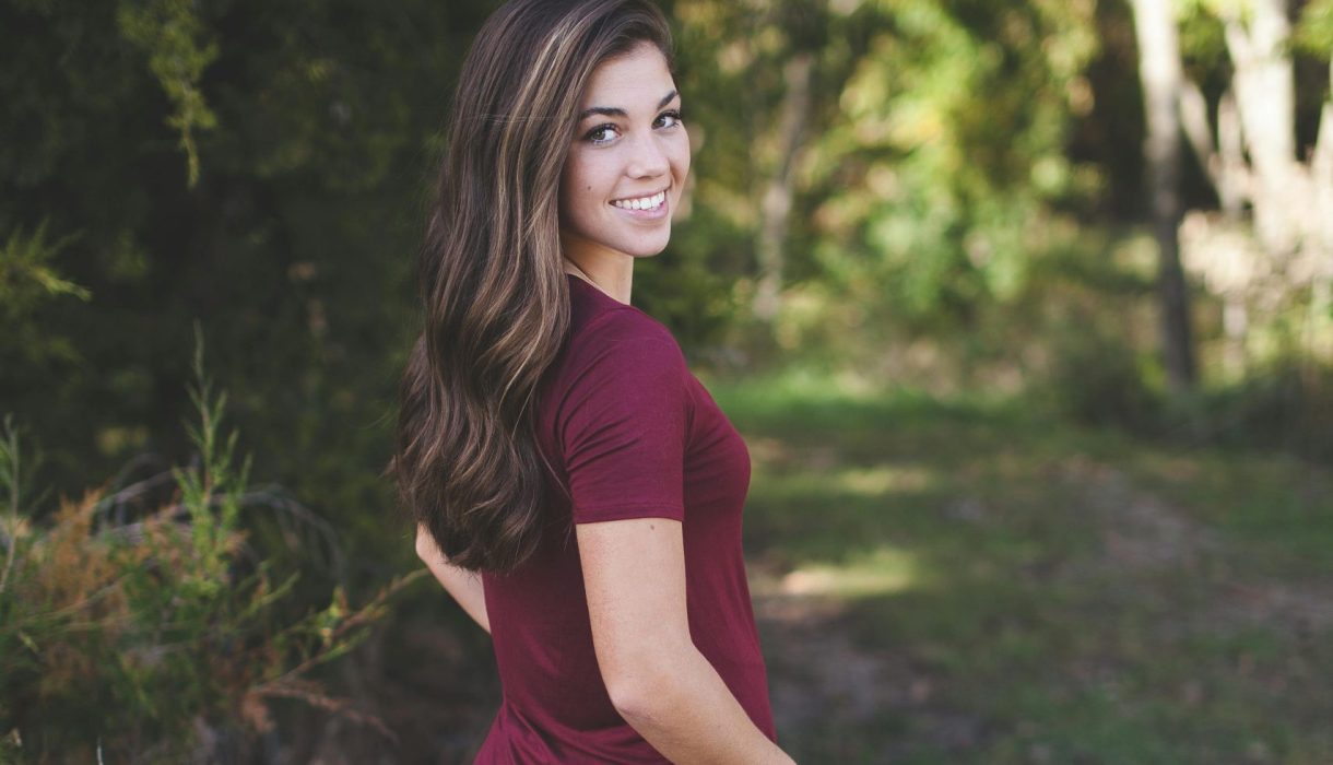 A young woman with long hair smiles outdoors in a forest setting, wearing casual attire.