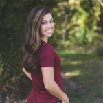 A young woman with long hair smiles outdoors in a forest setting, wearing casual attire.