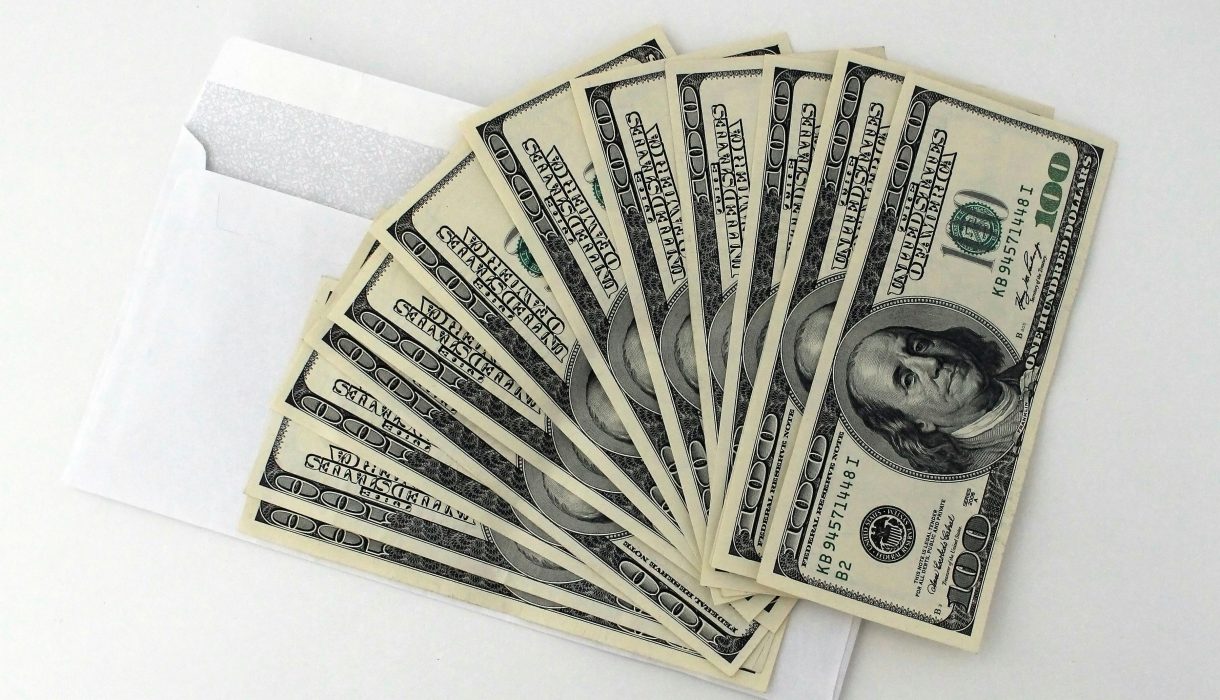 Fan of US $100 bills partially out of a white envelope on a white background.